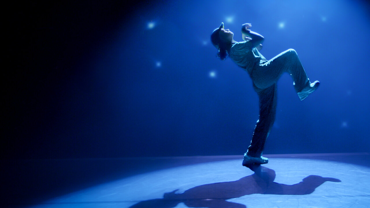 Raygun leans backward beneath blue stage lighting in a promotional still from Untold: Raygun — Breaking Badly.