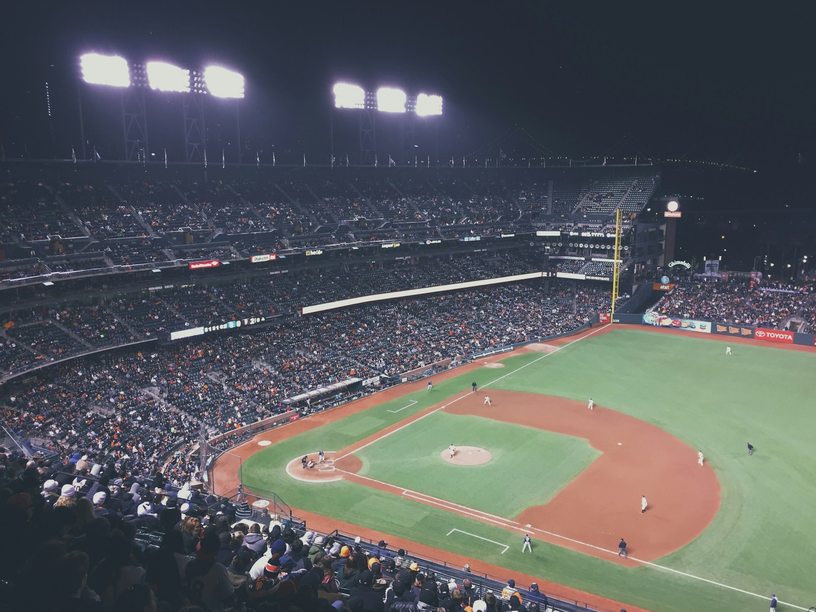 A crowded baseball stadium and its brightly lit diamond at night, seen from the upper stands.